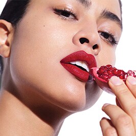 brunette woman with red lipstick holding a cut open pomegranate near her mouth